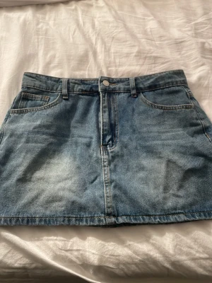 Blå jeanskjol med fickor - Säljer en klassisk blå jeanskjol med fem fickor och bälteshällor. Kjolen har en rak passform och stängs med knapp och dragkedja framtill. Perfekt att styla med en t-shirt eller hoodie för en chill look.