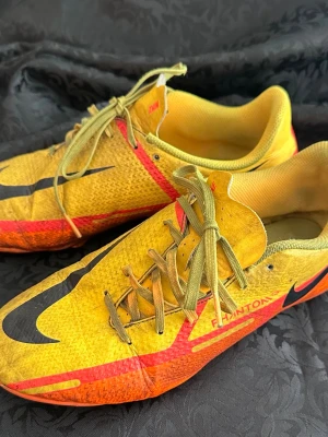 Nike Phantom GT2 gula fotbollsskor 37,5  - Säljer ett par Nike Phantom GT2 fotbollsskor i gult med svarta och röda detaljer. Skorna har snörning, låg profil och ovandel i syntetmaterial för bra bollkontroll. Swoosh-logga på sidan och tå, samt texten Phantom. Perfekta för dig som vill sticka ut på planen.