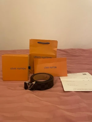 Louis Vuitton rutigt bälte i skinn - Säljer ett äkta Louis Vuitton bälte i mörkbrunt skinn med klassiskt rutigt mönster och silverfärgat LV-spänne. Bältet kommer med originalförpackning och kvitto, perfekt för dig som vill ha en lyxig accessoar med ikonisk design. 