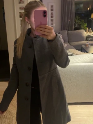 Mörkgrå klassisk ullkappa ifrån benetton - Snygg mörkgrå kappa med knappar framtill och figurnära passform. Ståkrage och diskreta sömmar i midjan ger en stilren siluett. Perfekt för kyligare dagar och enkel att matcha med olika outfits. Troligtvis i ullblandning för extra värme.
