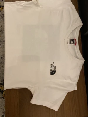 Vit t-shirt från The North Face - Vit t-shirt från The North Face med tryck på bröstet och stort grafiskt tryck på ryggen i blått och svart. Klassisk passform och rund halsringning. Storlek S