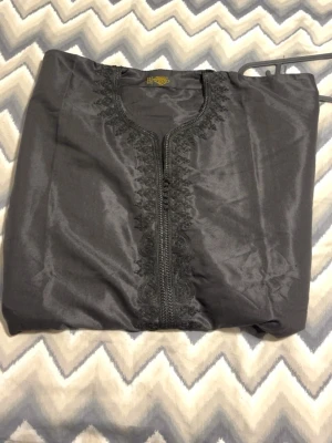 Svart kaftan med broderi - FINNS I BLÅ DE BARA SKIRVA !!!Säljer en svart kaftan med detaljerat broderi längs halsringningen och framtill. Materialet är glansigt och har en elegant finish. Perfekt för dig som vill ha något unikt och stilrent i garderoben.