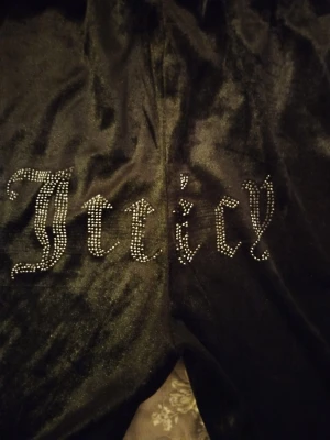 Svarta Juicy Couture mjukisbyxor - Säljer ett par svarta mjukisbyxor från Juicy Couture i glansigt velourmaterial. Byxorna har resår i midjan och texten 'Juicy' i strass på baksidan. Perfekta för en chill och trendig look.