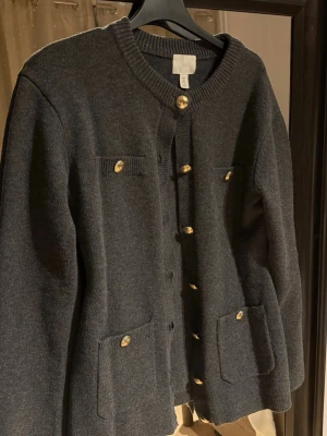 Mörkgrå kofta med guldfärgade knappar - Stilren mörkgrå kofta från H&M med rund halsringning och dekorativa guldfärgade knappar framtill. Koftan har två bröstfickor och två större fickor nedtill, alla med matchande knappar. Ribbstickade detaljer vid hals och ärmslut. Perfekt för lager på lager.