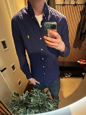 Marinblå skjorta från Ralph Lauren - Säljer en marinblå skjorta från Ralph Lauren med klassisk button-down krage och gul broderad logga på bröstet. Skjortan har långa ärmar, vita knappar och är tillverkad i bomull. Perfekt för en clean och stilren look.