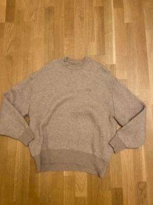 Beige Lacoste Stickad Tröja (Nyskick) - Säljer en riktigt snygg och stilren stickad tröja från Lacoste i en beige färg. Tröjan har den klassiska krokodilloggan broderad på bröstet i en diskret matchande färg (ton-i-ton), vilket ger ett exklusivt intryck. Tröjan är oanvänd utan prislapp, 10/10 skick. Storlek M. 