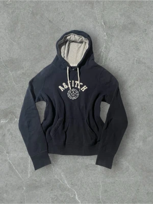 Mörkblå hoodie från Abercrombie & Fitch 40 - Mörkblå hoodie från Abercrombie & Fitch med broderad logga och text på bröstet. Hoodien har vit dragsko, mjuk insida och känguruficka framtill. Insidan av huvan är ljusgrå vilket ger en snygg kontrast. Perfekt för en avslappnad och sportig stil.