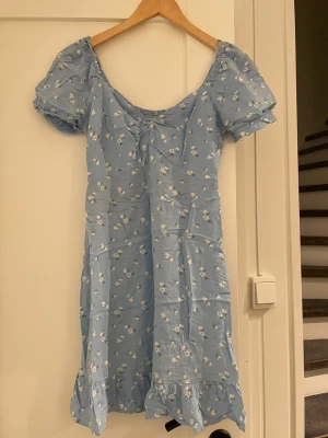 Blå blommig kortklänning med volang - Söt ljusblå kortklänning med små vita blommor, puffärmar och volangkant nertill. Klänningen har en lätt rynkad byst med knyt och smock i ryggen för skön passform. Perfekt för sommarens alla tillfällen!