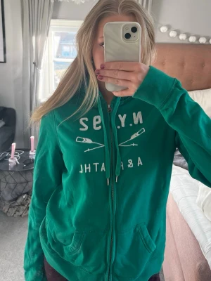 Grön hoodie från Abercrombie & Fitch - Snygg grön hoodie med dragkedja från Abercrombie & Fitch. Tryck på bröstet med texten N.Y. 92 och två korsade åror. Storlek L men jag själv brukar bära XS/S