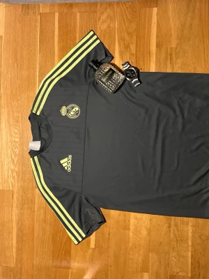 Real Madrid träningströja Adidas - Mörkgrå träningströja från Adidas med Real Madrid-logga på bröstet och gula detaljer. Tröjan har klassiska Adidas-ränder i gult längs axlarna och är tillverkad i ett lätt, snabbtorkande material. Perfekt för fotbollsträning eller gymmet.