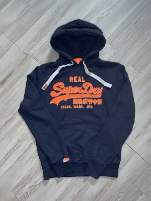 Mörkblå Superdry hoodie med tryck - Snygg mörkblå hoodie från Superdry med stor orange logga och japansk text på bröstet🤩 Längd 71cm         Bredd 57cm. storlek M kan även passa S 😇 skriv gärna vid snabbaffär samt funderingar går jättegärna ner vid snabbaffär🙌🏻
