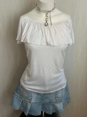 Vit offshoulder topp med volang - Snygg vit offshoulder topp med bred volang runt axlarna. Toppen är ärmlös och har en figurnära passform. Perfekt för varma dagar och enkel att styla med jeans eller kjol.