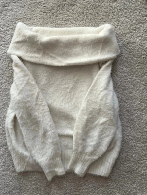 Vit off shoulder stickad tröja - Mysig vit stickad tröja med bred off shoulder-krage och långa ärmar. Tröjan har en fluffig och mjuk känsla och är väldigt bekväm. Passar dig som gillar en avslappnad men ändå trendig stil.