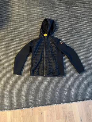 Svart vindjacka från Parajumpers - Snygg svart vindjacka från Parajumpers med huva och quiltade partier framtill och på sidorna. Jackan har dragkedja, fickor med dragkedja och en patch på ärmen. Materialmix med glansiga och matta ytor ger en sportig look.