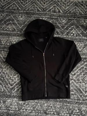 Svart zip hoodie från Jack And Jones  - Säljer en svart hoodie från Jack And Jones med dragkedja framtill och justerbar huva med snören. Hoodien har ribbade muddar vid ärmslut och nederkant samt en enkel, clean design. Tillverkad i mjukt bomullsmaterial för skön känsla. Helt skadefri och skriv vid eventuell frågor! Pris går att diskutera! 