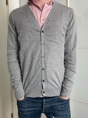 Grå cardigan i bomull, M - Snygg grå cardigan i Premium Cotton. Klassisk V-ringning, knäppning framtill med grå knappar och ribbade muddar vid ärmslut och nederkant. Perfekt att slänga över en skjorta för en clean och stilren look.