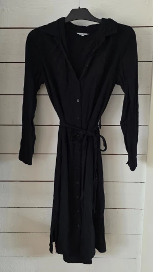 Svart långärmad klänning Bubbleroom - Svart långklänning från Bubbleroom med långa ärmar, knappar hela vägen framtill och ett matchande bälte i midjan. Klänningen har klassisk krage och är gjord i ett mjukt tyg som ger en avslappnad men stilren look.