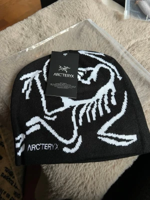 Svart mössa från Arc'teryx - Snygg svart mössa från Arc'teryx med vit grafisk logga och text på framsidan. Tillverkad i mjukt, stretchigt material som sitter skönt på huvudet. Perfekt för dig som vill ha en stilren och sportig look.