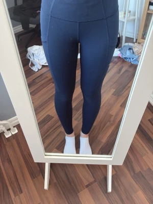 Mörkblå leggings med fickor - Säljer ett par mörkblå leggings med hög midja och praktiska sidofickor. De har en tight passform och är tillverkade i ett stretchigt material som sitter skönt på kroppen. Liten vit logga baktill i midjan för en sportig look. (Pris går att diskutera)