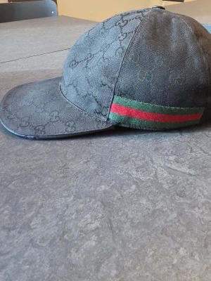 Gucci keps svart (äkta) - Snygg svart keps från Gucci med klassiskt GG-monogram över hela kepsen. På sidan finns en grön och röd rand som ger den en ikonisk look. Justerbar rem baktill i läder och böjd skärm. Tillverkad i slitstarkt tygmaterial.
