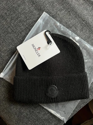 Svart Moncler mössa i ull - Säljer en svart ribbstickad mössa från Moncler med uppvikt kant och Moncler-logga framtill. Mössan är tillverkad i mjuk ull och har en klassisk, stilren design som passar perfekt till höst och vinter.