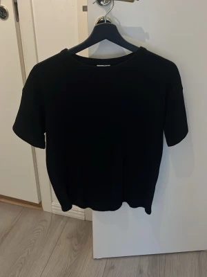 Svart stickad t-shirt från Kappahl - En enkel svart t-shirt från Kappahl med rund halsringning och korta ärmar. Perfekt basplagg som funkar till allt och har en klassisk passform. Tillverkad i mjuk bomull för skön känsla hela dagen.