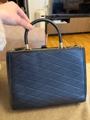 Svart quiltad handväska med gulddetaljer - Snygg svart handväska i quiltat mönster med guldfärgade detaljer och dragkedja upptill. Väskan har både handtag och avtagbar axelrem för olika stilar. Tillverkad i konstläder och har en stilren, fyrkantig form som passar till många outfits.