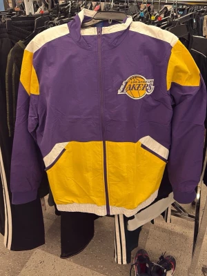 Lakers vindjacka i lila och gul - Storlek XL men funkar perfekt för L skulle jag säga. Kobes kända uppvärmningsjacka. Snygg vindjacka med Los Angeles Lakers-logga, lila och gula färgblock samt vita detaljer. Jackan har dragkedja, hög krage och två fickor framtill. Perfekt för basketfans som vill sticka ut. Materialet är lätt och vindtätt. 