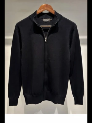 Svart kofta med dragkedja Dressmann storlek S - Svart kofta från Dressmann i Supima bomull, storlek S. Koftan har hel dragkedja framtill och ribbad krage. Perfekt för dig som gillar stilrena och enkla plagg med snygg passform. Passar till många olika outfits och är riktigt skön att ha på sig.