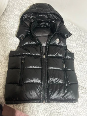 Svart dunväst från Moncler med huva - Snygg svart dunväst från Moncler med glansig finish och avtagbar huva. Västen har två dragkedjeförsedda fickor framtill och klassisk Moncler-logga på bröstet. Perfekt för lager på lager och streetwear-vibe. Materialet är dunfyllning och yttertyget är blankt.