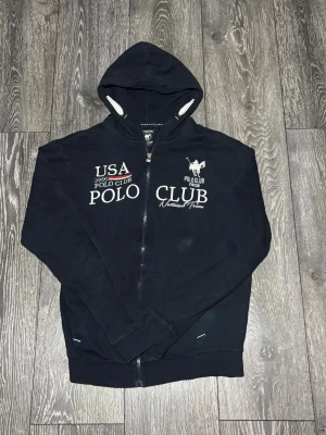 Marinblå hoodie från Polo Club Vinson - Snygg marinblå hoodie med dragkedja från Polo Club Vinson. Tryck på bröstet med texten 'USA Polo Club' och logga. Mjuk insida, två fickor fram och vit snörning i huvan. Perfekt för en avslappnad och sportig stil.