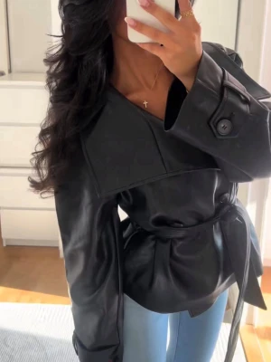 Svart oversized skinnjacka med bälte - Snygg svart skinnjacka från Gina tricot med bred krage och knytbälte i midjan. Jackan har stora dekorativa knappar vid ärmsluten och en cool, modern look. Perfekt för dig som vill ha en trendig och edgy stil.