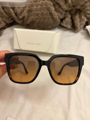 Svarta fyrkantiga solglasögon Michael Kors - Stilrena solglasögon från Michael Kors med svart, fyrkantig båge och bruna tonade glas. Snygg guldfärgad MK-logga på sidorna ger en lyxig känsla. Perfekta för att lyfta din look under soliga dagar.