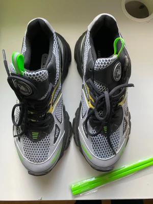 Axel arigato x Caliroots sneaker   - Chunky sneakers från Aircross i grått, svart och vitt meshmaterial med detaljer i neonlime och gult. Skorna har tjock sula, svarta snören och gröna dragflikar. Sportig design med ventilerande mesh och robust känsla.