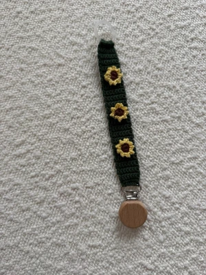 Virkad napphållare med solrosor - Handgjord virkad napphållare i mörkgrönt garn med tre gula solrosor som dekoration. Fästet är i trä och metall, och öglan upptill är i plast. Perfekt accessoar för att hålla koll på nappen och ge en personlig touch.