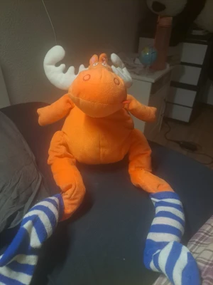 Orange mjukisälg med randiga ben - Säljer en mjukisälg i orange plysch med vita horn och långa ben i blåvitrandigt tyg. Ansiktet har broderade ögon och nos. Perfekt som dekoration eller gosedjur för dig som gillar unika mjukdjur.