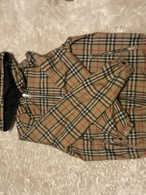 Reversible vindkacka burberry - Snygg vindjacka från Burberry med klassiskt beige rutmönster och svarta, röda och vita detaljer. Jackan är vändbar med en sida i helsvart och har dragkedja framtill samt fickor med dragkedja. Tillverkad i lätt, glansigt material och har elastiska muddar vid ärmslut och nederkant.