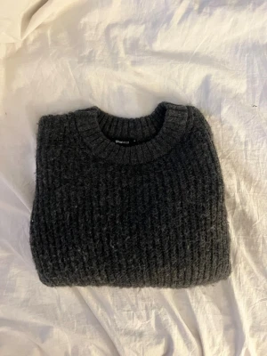 Mörkgrå stickad tröja från Gina Tricot - Mysig mörkgrå stickad tröja från Gina Tricot med grov ribbstickning. Tröjan har rund halsringning, långa ärmar och breda muddar. Enkel att matcha med jeans eller kjol.