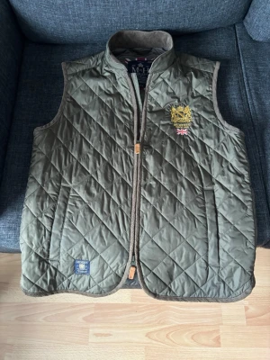 Olivgrön quiltad dunväst från Morris - Snygg olivgrön dunväst från Morris med quiltat mönster och guldfärgat broderi på bröstet samt en liten brittisk flagga. Perfekt nu när det börjar bli lite varmare 