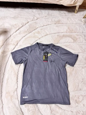 träningströja Under Armour - Snygg grå kortärmad träningströja från Under Armour med diskret mönster och logga på bröstet. Tillverkad i ett lätt och snabbtorkande funktionsmaterial som passar perfekt till löpning eller gym. Rund halsringning och bekväm passform.