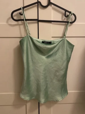 Mintgrön satinlinnetopp från Gina Tricot - Säljer en mintgrön linnetopp från Gina Tricot i glansigt satinliknande material. Toppen har tunna axelband och en lös, något utsvängd passform. Perfekt för dig som gillar en enkel men ändå trendig look.