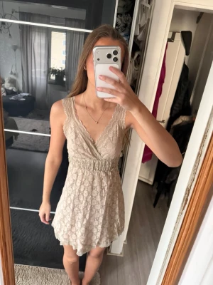 Beige spetsklänning med v-ringning - Söt kort klänning i beige spets med omlott och v-ringning. Klänningen är ärmlös och har en markerad midja med volangkant nertill. Perfekt för sommarens alla tillfällen.
