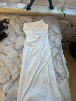 Vit axelbandslös långklänning - Säljer en vit siden axelbandslös långklänning i glansigt material. Klänningen har en draperad överdel och en slits framtill som ger en snygg touch. Perfekt för student eller ett festligare tillfälle.Den är ifrån Asos och är i storlek Xs/34