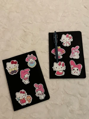 Svarta anteckningsböcker med Hello Kitty - Två svarta anteckningsböcker dekorerade med färgglada Hello Kitty- och My Melody-klistermärken i rosa, vitt och rött. Perfekta för skolarbete, dagbok eller kreativa anteckningar. Omslaget är mjukt och böckerna har ett stilrent, lekfullt utseende.