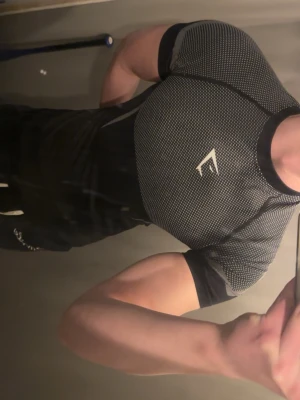 Svart tight träningströja Gymshark - Säljer en svart tight träningströja från Gymshark med grått mönster och logga på bröstet. Tröjan har korta ärmar och är tillverkad i ett stretchigt funktionsmaterial som andas, perfekt för gym eller löpning.
