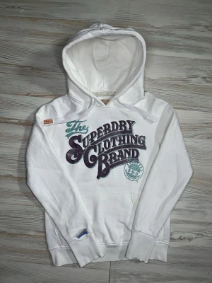 Vit superdry hoodie - Vit hoodie från Superdry med stort lila och ljusblå tryck på bröstet där det står ’The Superdry Clothing Brand’🤩 Hoodien är svårt och få tag på och är i bra skick🙌🏻 Hör av er vid funderingar samt snabbaffär😇