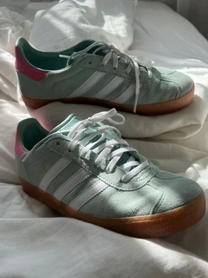 Adidas Gazelle sneaker - Snygga Adidas Gazelle sneakers i mintgrönt mocka med klassiska vita ränder och gummisula. Skon har vita skosnören, rosa detalj på hälen och guldfärgad Gazelle-text på sidan. Perfekt till våren! Stl 38, normal. Använd fåtal gånger inga tydlige defekter nästan nyskick 💘