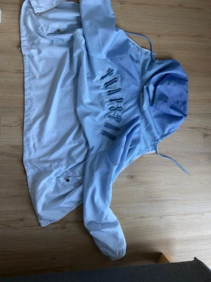 Ljusblå vindjacka med huva - Snygg ljusblå vindjacka med huva och dragsko. Jackan har unika broderade detaljer i form av bokstäver på ryggen i en mörkare blå nyans. Lätt material, perfekt för blåsiga dagar. Stängs med tryckknappar och har en avslappnad passform.