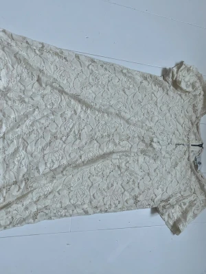 Vit spetsklänning med kort ärm - Söt vit spetsklänning med korta ärmar och rund hals. Klänningen har ett vackert blommigt spetsmönster och är i en kort modell. Perfekt för sommarens alla tillfällen. Dragkedja i ryggen för enkel påklädning.