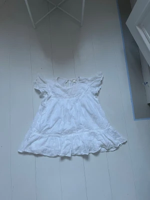 Vit volangblus med spetsdetaljer - Söt vit blus med volangkant och spetsdetaljer. Blusen har rund halsringning, korta volangärmar och knäppning bak i nacken. Tillverkad i ett tunt, luftigt tyg som ger en romantisk känsla. Perfekt att styla med jeans eller kjol för en fräsch look.